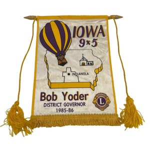 Può includere: Uno stendardo giallo e bianco con una mongolfiera viola e gialla, una mappa dell'Iowa e il testo "Iowa 9x5 Indianola Bob Yoder District Governor 1985-86 Lions International".