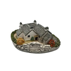 May include: A miniature stone cottage with a gray slate roof and white walls. The cottage is surrounded by a stone wall and green foliage. The base of the cottage has the word "Heritage" and a copyright symbol.