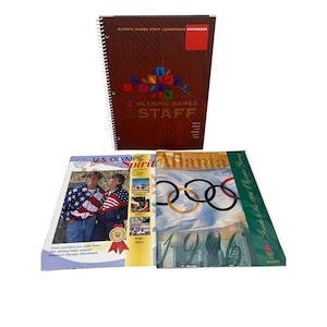 May include: A selection of Olympic Games publications, including a spiral-bound handbook with "OLYMPIC GAMES STAFF LEADERSHIP HANDBOOK" on the cover. Other publications include "U.S. Olympic Spirit" and "Atlanta" with Olympic rings.
