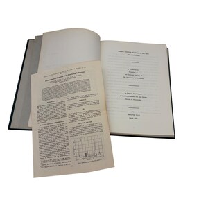 Pode incluir: Um livro aberto com capa preta, exibindo várias páginas de texto e diagramas. As páginas incluem uma página de título e um artigo de pesquisa intitulado "Antiferromagnetic Properties of the Iron Group Trifluorides". O livro está aberto, revelando conteúdo detalhado.