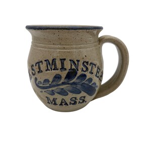 May include: A beige ceramic mug with a dark blue rim and handle. The mug features the text "STMINSTER MASS." in dark blue lettering, along with a blue leaf design. The mug has a rustic, handcrafted appearance.
