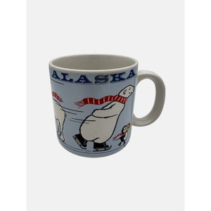 May include: A white ceramic mug with a blue background and a cartoon image of a polar bear wearing a red and white scarf ice skating. The text "ALASKA" is printed in blue on the mug.