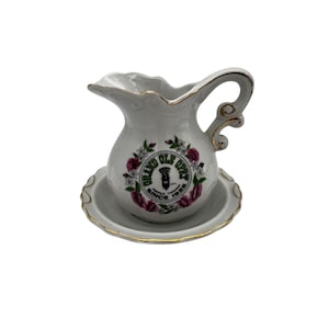 Puede incluir: Un juego de jarra y platillo de porcelana blanca con ribete dorado. La jarra presenta un logotipo verde y blanco que dice "Grand Ole Opry" con las palabras "Nashville, Tennessee" y "Since 1925" debajo. El logotipo está rodeado de flores rosas y blancas.