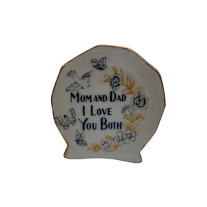 Puede incluir: Un plato de cerámica blanca con borde festoneado y ribete dorado. El plato presenta diseños de palomas, mariposas y flores en negro y dorado. El texto "Mom and Dad I Love You Both" está escrito en azul marino.