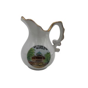 May include: White ceramic pitcher with a gold rim and handle. The pitcher features a colorful image of a building with the text "SIX FLAGS OVER GEORGIA". The pitcher is a decorative item.