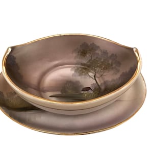 Puede incluir: Un juego de dos cuencos de cerámica con una escena de paisaje pintada a mano. El cuenco es de color marrón claro con un borde dorado y el platillo es de color gris claro con un borde dorado. La escena representa una pequeña casa, árboles y un río.