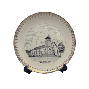 Peut inclure: Une assiette décorative vintage avec une illustration en noir et blanc de l'église méthodiste Ebenezer à Calhoun, Tennessee. L'assiette a un fond crème avec une bordure florale dorée. L'illustration de l'église est détaillée, sur fond de ciel nuageux.