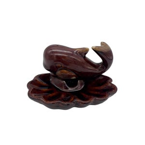 May include: A brown ceramic whale figurine with a glossy finish. The whale is perched on a matching brown, scalloped-edge dish. The figurine has a detailed design, with visible eyes and fins. The overall aesthetic is vintage and decorative.