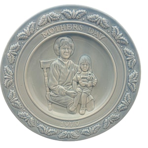 May include: A blue ceramic plate with a raised image of a mother and child. The plate has a white border with a leafy design and the words "Mother's Day 1979" in the center.