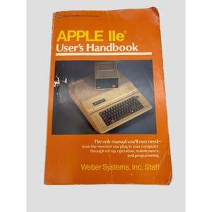 May include: An orange Apple IIe User's Handbook with yellow text. The cover features an illustration of an Apple IIe computer and a disk drive. The text reads: The only manual you'll ever need—from the moment you plug in your computer.