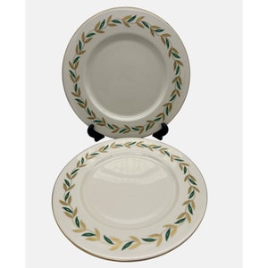 Può includere: Due piatti in ceramica bianca con bordo dorato e bordo decorativo di foglie verdi e dorate. I piatti sono esposti su un supporto nero su sfondo bianco. I piatti sono probabilmente per mangiare o esporre.
