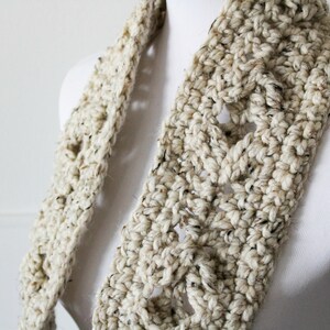 May include: A thick, hand-crocheted scarf in cream with black flecks. The scarf has an open weave design, creating a textured look. It is displayed on a white mannequin. The scarf is a cozy and stylish accessory for colder weather.