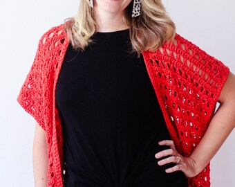May include: A woman wearing a red crochet shawl over a black top and blue jeans. The shawl has an open, airy design. The woman is smiling and wearing earrings.