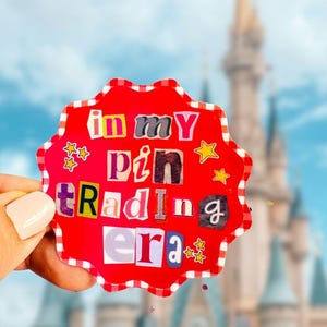May include: A red, scalloped-edge sticker with the text "in my pin trading era" in a collage style. The background features a blurred image of a castle under a blue sky.