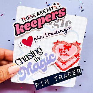May include: A collection of colorful stickers with phrases like "These are my keepers" and "Chasing the Magic." The stickers feature hearts, stars, and a heart-shaped design with the words "Pin Trader." The stickers are on a white card.