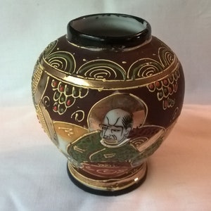 May include: A small, round, brown ceramic vase with gold accents and a painted image of a man with a beard. The vase has a traditional Japanese design.