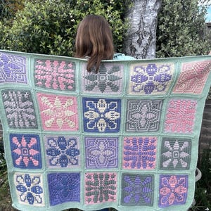 May include: A handmade crochet blanket with a patchwork design. It features floral patterns in pink, purple, blue, and grey, with a light green border. The blanket is displayed outdoors against a natural backdrop.