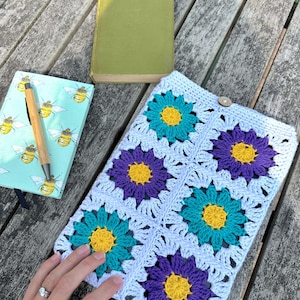 May include: A rectangular crochet sleeve featuring a light blue base with four large flower motifs in alternating teal and purple petals with yellow centers. The sleeve is secured with a small wooden button at the top edge and has a textured finish.