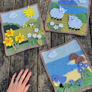 May include: Three crocheted squares with countryside scenes. One shows a sun, flowers, and bees. Another depicts sheep in a field. The third features birds and mushrooms. Each square has a brown border, set against a wooden surface.