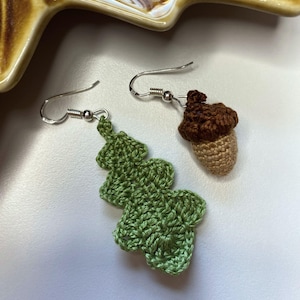 May include: A pair of handmade crochet earrings. One earring is a green leaf shape, and the other is a brown and tan acorn. Both earrings have silver-colored fish hook ear wires. The earrings are on a white surface.