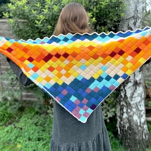 Op de afbeelding: Een kleurrijke, handgehaakte sjaal met een geometrisch patroon. De sjaal heeft een levendig kleurenpalet, waaronder oranje, geel, blauw en paars. De driehoekige sjaal heeft een geschulpte rand. De sjaal wordt voor een persoon gehouden.
