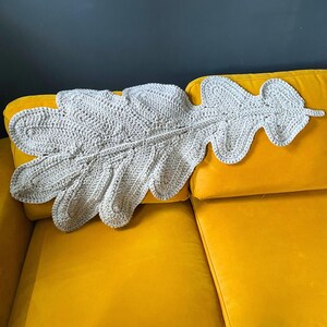 May include: A large, beige crocheted leaf-shaped blanket rests on a bright yellow sofa. The blanket's intricate stitchwork creates a textured surface. The sofa's vibrant color contrasts with the neutral tones of the blanket, creating a cozy and inviting aesthetic.
