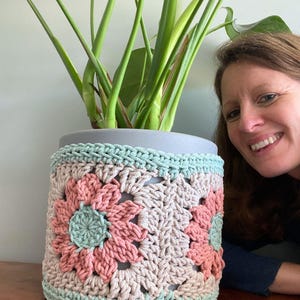 May include: A grey ceramic plant pot with a crocheted cosy. The cosy features a floral design in pink, beige, and mint green. The plant pot holds a green leafy plant.