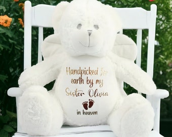May include: White plush teddy bear with angel wings, sitting on a white rocking chair. The bear has the text "Handpicked for earth by my Sister Olivia in heaven" embroidered on its chest, with a pair of brown footprints below the text.