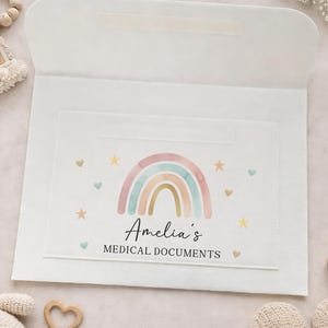 May include: A white medical document holder with a rainbow design in pastel colors. The holder features the text "Amelia's MEDICAL DOCUMENTS" and is decorated with hearts and stars. The holder is open, revealing the interior.