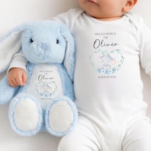 May include: A light blue plush bunny and a white baby onesie. The bunny and onesie both feature the text "Hello World I'm Oliver" with an elephant graphic and the year "Born in 2026". The onesie has long sleeves.