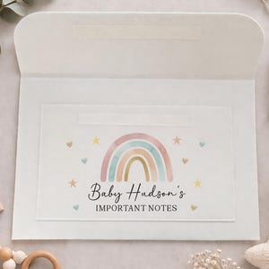 May include: A white envelope with a clear window featuring a watercolor rainbow design and the text "Baby Hudson's IMPORTANT NOTES." The design includes pastel colors, stars, and hearts. The envelope is open, revealing the interior.