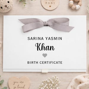 May include: A white birth certificate box with a gray ribbon, the name "Sarina Yasmin Khan" and the words "Birth Certificate" are printed on the front. The box is surrounded by baby-themed decorations, including a wooden star, a wooden disc that says "hello baby", and a stuffed bunny.