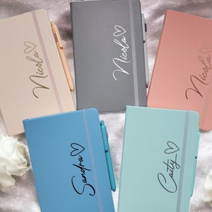 May include: Five personalised notebooks with a heart design and a pen. The notebooks are in different colours: pink, grey, light blue, and blue. The names on the notebooks are Nicola, Sandra, and Cathy.