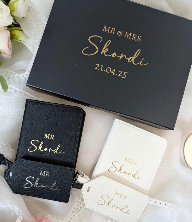 Op de afbeelding: Een zwarte geschenkdoos met een gouden inscriptie "MR & MRS Skordi 21.04.25". Twee paspoorthouders, een zwarte en een witte, met bijpassende bagagelabeltjes. De paspoorthouders en labeltjes zijn gepersonaliseerd met de naam "Skordi" in gouden letters.