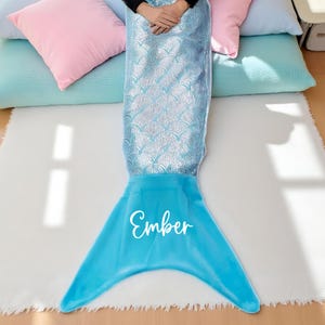 May include: A mermaid tail blanket featuring a silver scale pattern on the upper section and a solid bright blue fin at the bottom. The name Ember is printed in white script lettering across the centre of the blue fin section of the blanket.