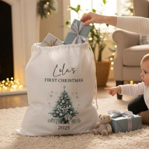 May include: A white Christmas gift bag with a drawstring closure, featuring the text "Lola's FIRST CHRISTMAS" and a Christmas tree design. The bag is filled with gifts, and a baby is pointing at a gift on the floor.