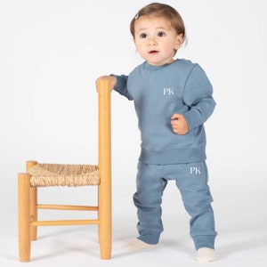 May include: A toddler wearing a blue sweatshirt and matching sweatpants with the letters "PK" embroidered on the front and back. The child is standing next to a small wooden chair.