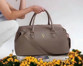May include: A taupe-colored travel bag with a long shoulder strap and two top handles. The bag has a gold zipper and a gold monogram with the initials "NT" and a heart.