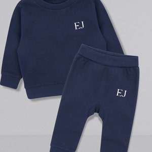 May include: A navy blue two-piece baby outfit with long sleeves and pants. The top has a round neckline and the pants have an elastic waistband. Both pieces have the initials "EJ" embroidered on the front in white.