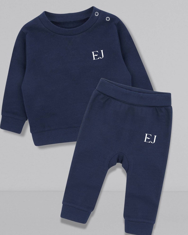May include: A navy blue two-piece baby outfit with long sleeves and pants. The top has a round neckline and the pants have an elastic waistband. Both pieces have the initials "EJ" embroidered on the front in white.