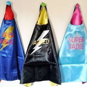 May include: Three superhero capes with personalized names. The blue cape says "Super Albert" with a yellow lightning bolt. The black cape says "Alfred" with a white lightning bolt. The light blue cape says "Super Sadie" with a silver star.