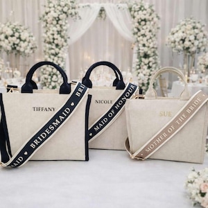 May include: Three beige tote bags with black handles and straps. The bags have text that reads "Bridesmaid", "Maid of Honour", and "Mother of the Bride". The bags are on a white surface with a floral background.
