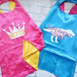 May include: Two superhero capes, one pink with a white crown and the name "Hettie" and one blue with a white dinosaur silhouette and the text "Superhero Sienna".