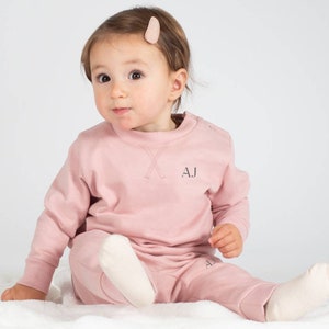 May include: A pink baby sweatshirt and sweatpants set with the initials "AJ" embroidered on the front. The set is made of soft, comfortable fabric and is perfect for everyday wear.