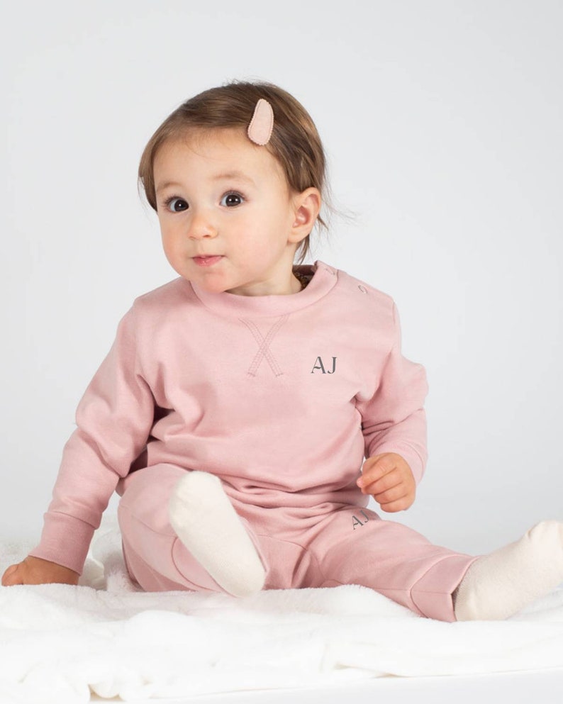 May include: A pink baby sweatshirt and sweatpants set with the initials "AJ" embroidered on the front. The set is made of soft, comfortable fabric and is perfect for everyday wear.