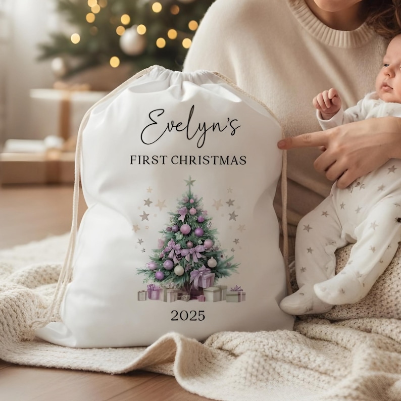 May include: White drawstring gift bag with the text "Evelyn's FIRST CHRISTMAS" and the year 2025. The bag features a watercolor illustration of a Christmas tree decorated with purple ornaments and gifts. The bag is held by a person holding a baby.