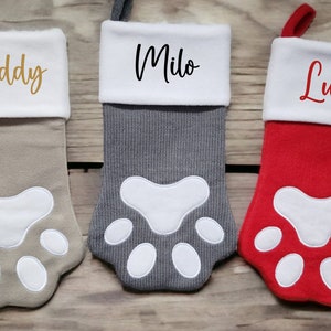 May include: Three pet-themed Christmas stockings in beige, grey, and red, each with a white paw print design. The names "Buddy", "Milo", and "Luna" are embroidered on the white cuff. The stockings are designed to look like a pet's paw.