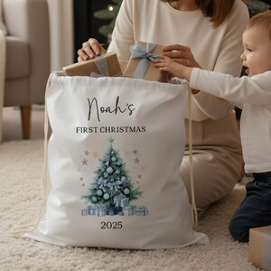 May include: A white drawstring Christmas gift bag with the text "Noah's First Christmas 2025". The bag features a watercolor Christmas tree design with blue ornaments and presents. Two wrapped gifts are being placed inside the bag.