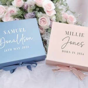 May include: Two personalised gift boxes, one blue with white text and a blue ribbon, and one pink with grey text and a pink ribbon. The blue box says "Samuel Donaldson 24th May 2024" and the pink box says "Millie Jones Born in 2024".