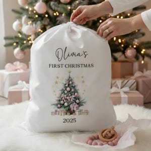 May include: A white Christmas gift bag with a drawstring closure. The bag features the text "Olivia's FIRST CHRISTMAS" above a Christmas tree design with pink ornaments and bows. The year "2025" is printed below the tree.